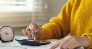 A woman in a yellow jumper does tax calculations at a desk with a calculator