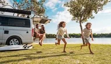 Two children run along a lakeside while their parents watch from the back of a campervan