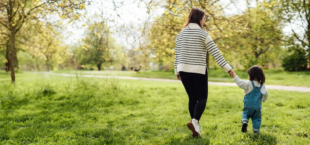 Woman walks across a grassy part of a park on an autumn afternoon while holding her young daughter's hands.