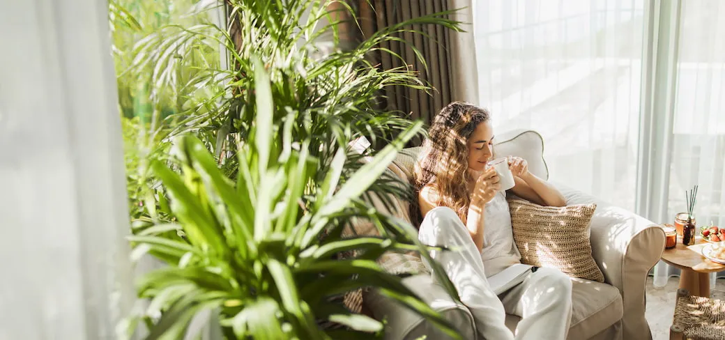 Woman sipping a cup of coffee while sitting in a lounge chair with light filtering through indoor plants.