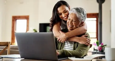 Woman hugs her grandmother as they sit in front of a laptop.