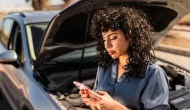 A young woman uses her mobile phone to contact Roadside Assistance in front of her broken-down car on the side of the road.