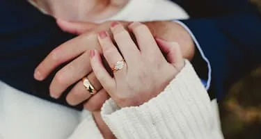 A diamond engagement ring sits on a person's finger while holding their partner's hand