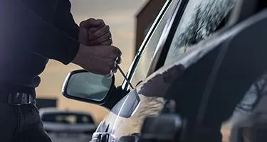 A man wearing black clothing using a poker / slim jim to break into the front passenger door of a car.
