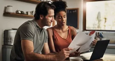 Young couple sit in kitchen looking at paperwork in front of a laptop.