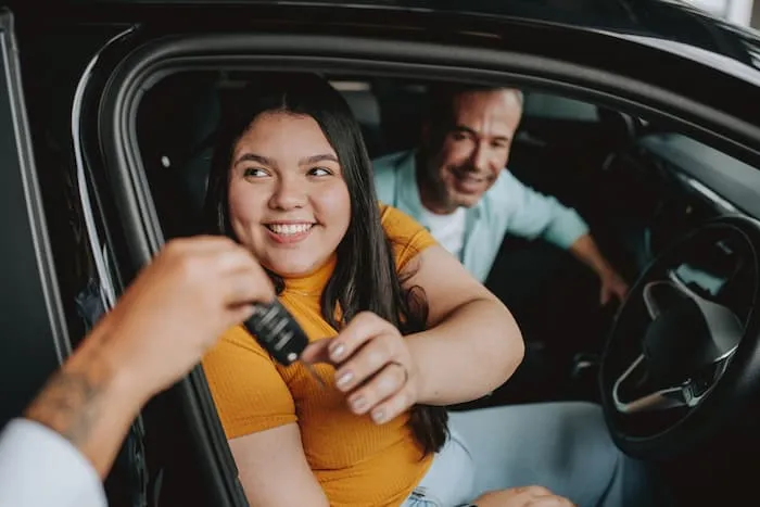 Daughter sits next to her father as she reaches for new car keys in a car she purchased from a dealership.