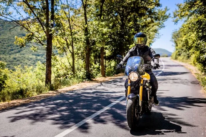 A motorcyclist rides a standard motorcycle on a paved mountain road on a sunny day.