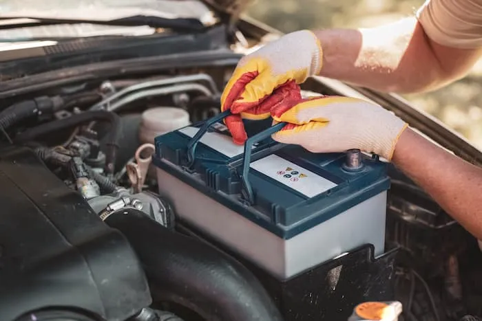 A mechanic wearing gloves removes a car battery from under the hood of a car.