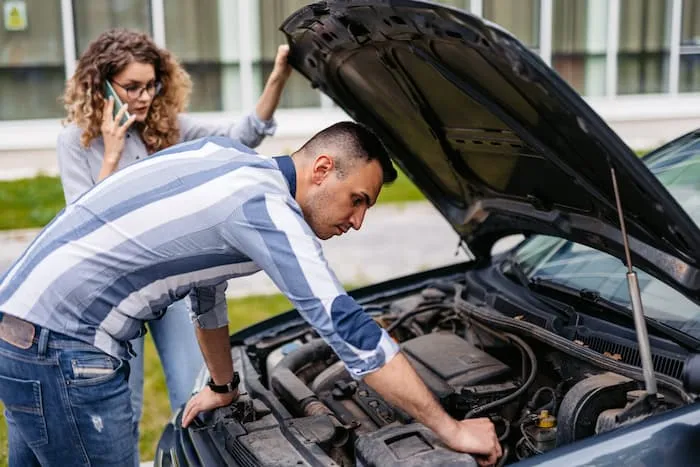 A woman calls roadside assistance for help with a car breakdown while a man looks under the hood of the car.