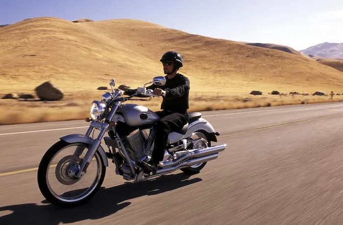 A man rides a shiny silver cruiser motorcycle on a road with rolling hills in the background.