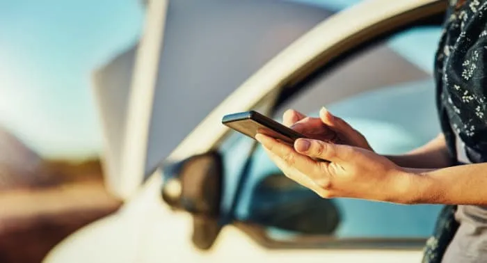 A woman looks down at her phone beside her broken down car