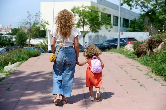 A mother holds her daughters hand as they walk down a footpath near traffic on the road.
