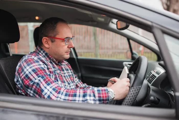 A driver wearing glasses takes notes on the number of kilometres he has driven while sitting inside his car.