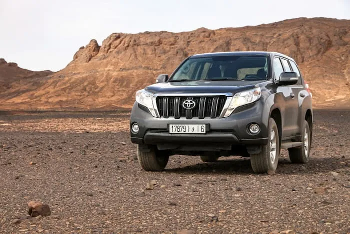 grey Toyota Land Cruiser Prado parked on gravel road in desert in front of rocks