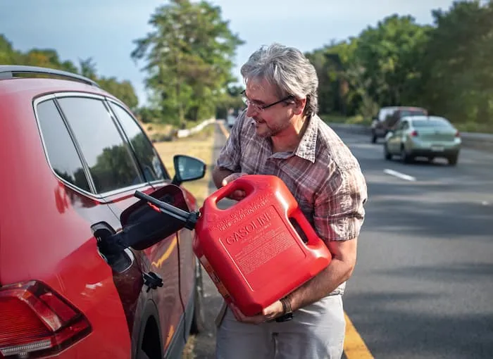 An older man refills his car's petrol tank from a red plastic container on the side of the road.