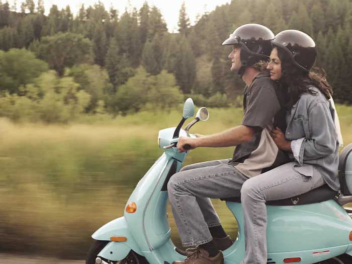 A young couple ride a pale blue scooter through the countryside.