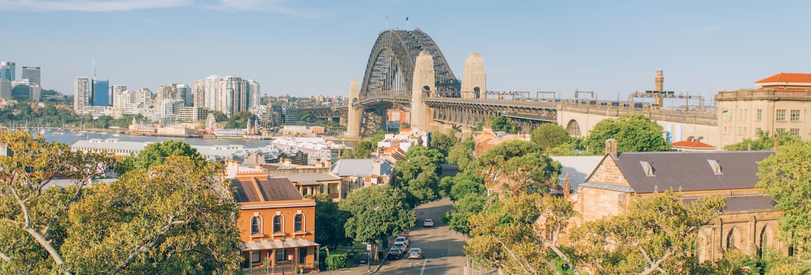 Green trees frame streets that wind through old brick houses underneath the Sydney Harbour Bridge in Sydney, New South Wales.