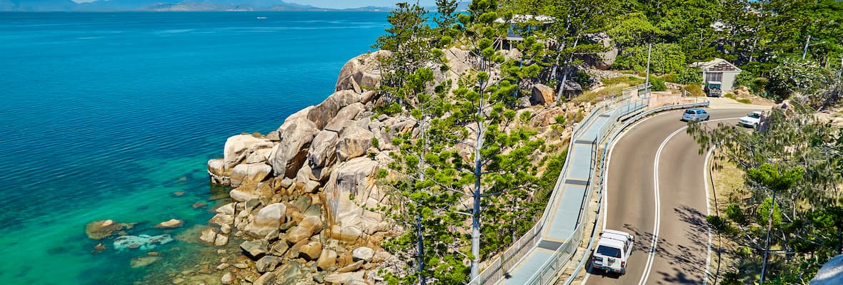 An elevated roadway winds through rocky cliffsides, green trees and blue ocean water at Geoffrey Bay on Magnetic Island.