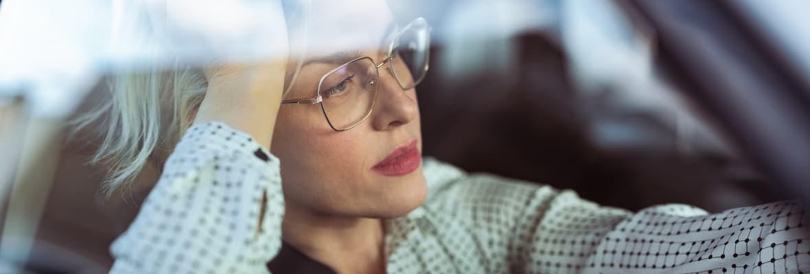 A tired woman wearing glasses sits in her car during a traffic jam.