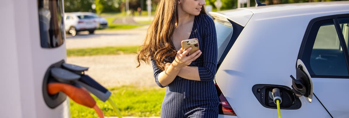 Woman leaning on car checks phone while electric vehicle charges
