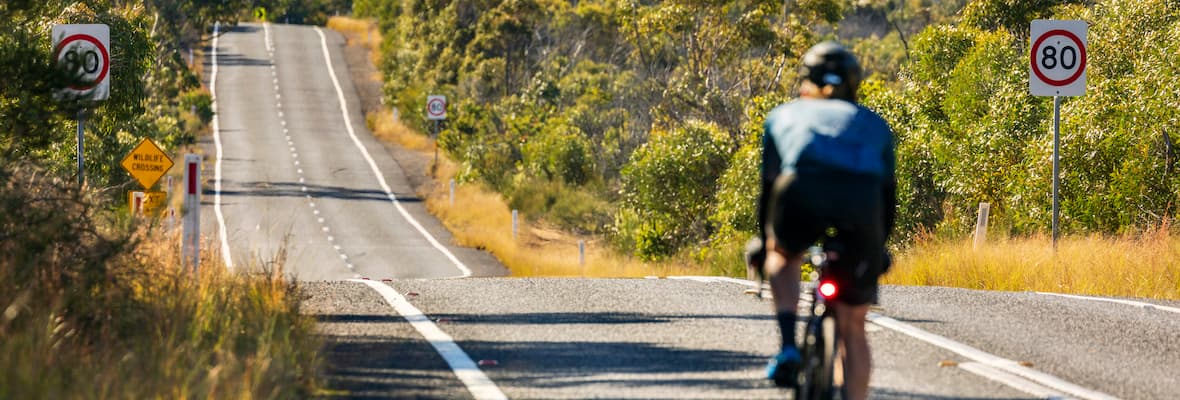 A cyclist rides downhill, passing green bushland on the road towards Sydney, New South Wales.