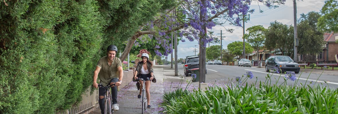 A young couple cycles underneath a Jacaranda tree on a footpath in suburban Sydney.