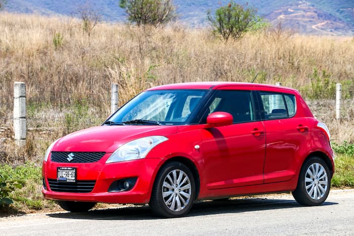 Red Suzuki Swift is parked on the side of the road next to dead grass