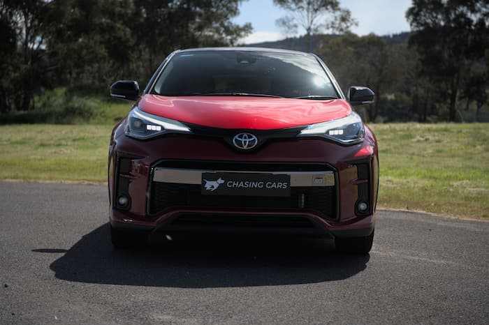 Red toyota C-HR parked on road outside with headlights on in front of dead grass