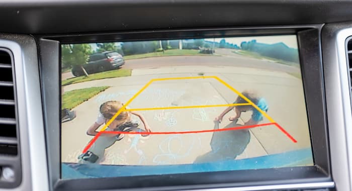 A rear view camera screen shows children playing behind a car
