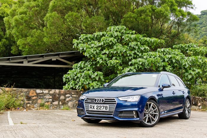 Blue Audi A4 parked in front of a brick fence and green leaves on a tree