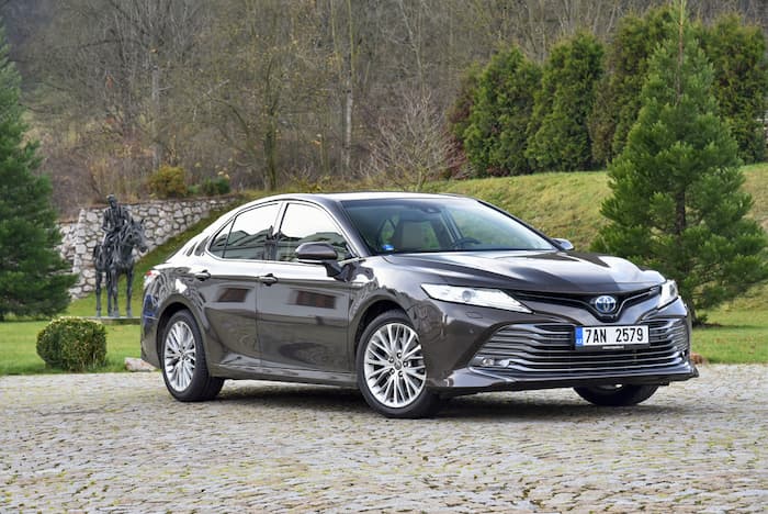 Grey Toyota Camry is parked on a stone driveway with green trees in the background