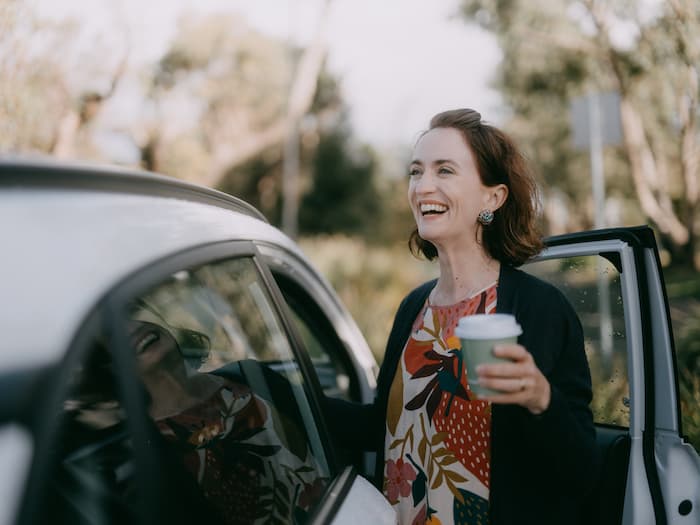 A happy woman stands in between her car and her open car door as she holds a coffee.