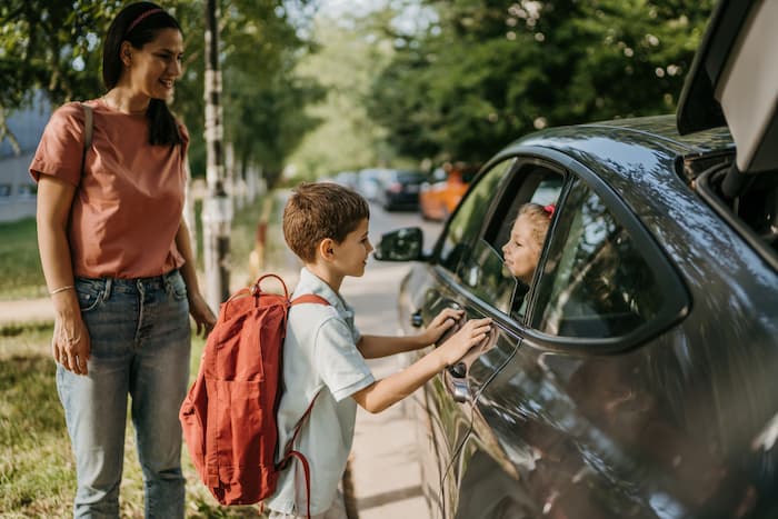 A young boy says goodbye to his sister in the car before his mother drops him off at school.