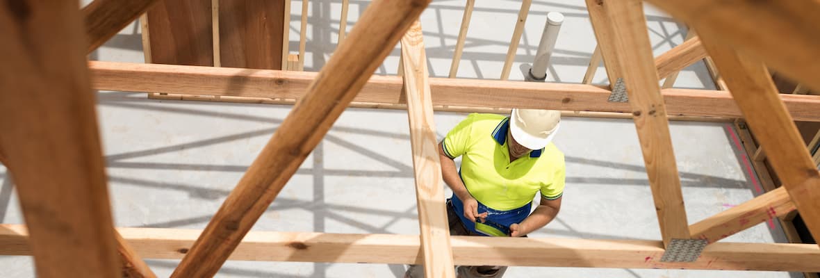 A builder checks a building plan on a tablet under the wooden frame of a newly built home.