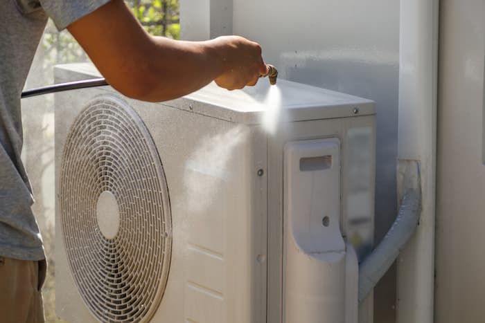 A man uses a hose to spray water onto an outdoor air conditioner unit.
