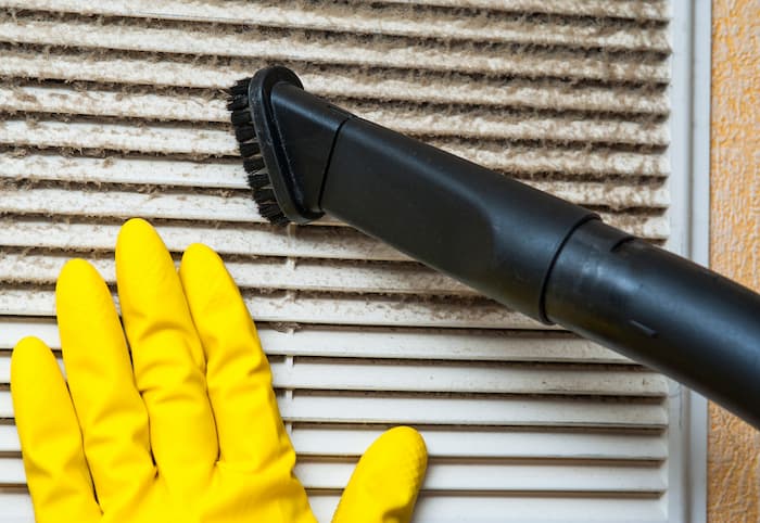 A person wearing yellow gloves uses a soft brush to gently remove dust from air conditioner fins.