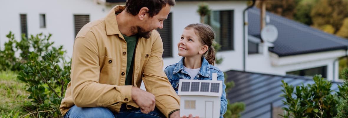 A dad shows his daughter a paper model of a house with solar panels, crouched down on grass in front of the real house. 