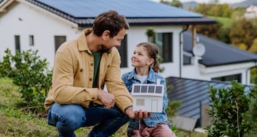 A dad shows his daughter a paper model of a house with solar panels, crouched down on grass in front of the real house. 