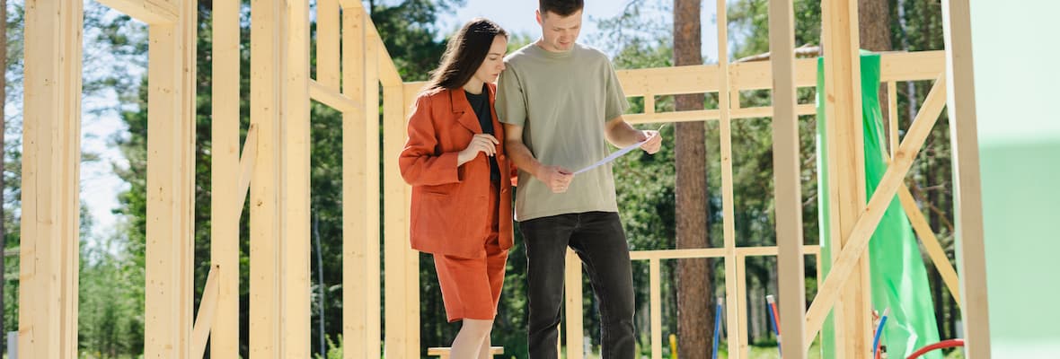 A young couple reviews building plans standing inside the wooden frame of their home. 