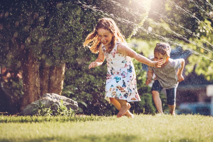 young boy and girl running through sprinkler on grass