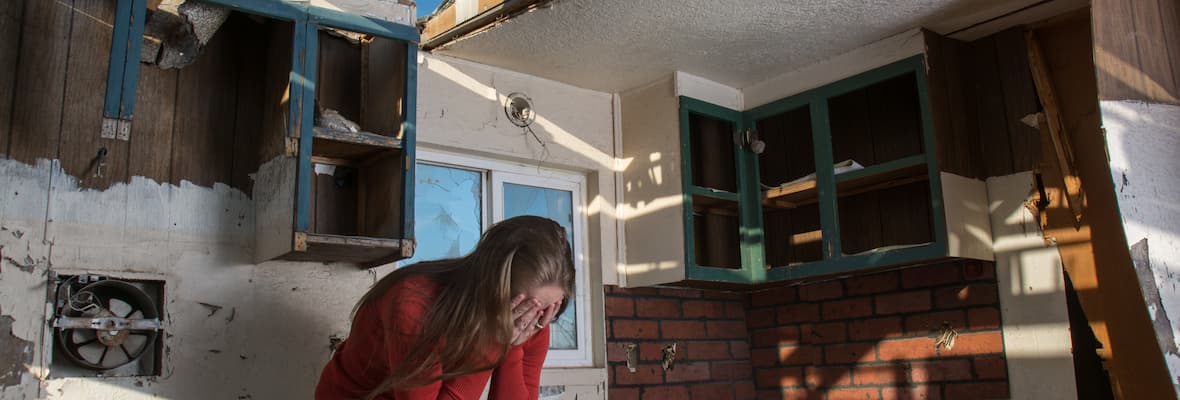 A woman sits on a kitchen bench with her head in her hands, in the middle of her damaged home. 