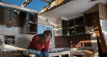 A woman sits on a kitchen bench with her head in her hands, in the middle of her damaged home. 