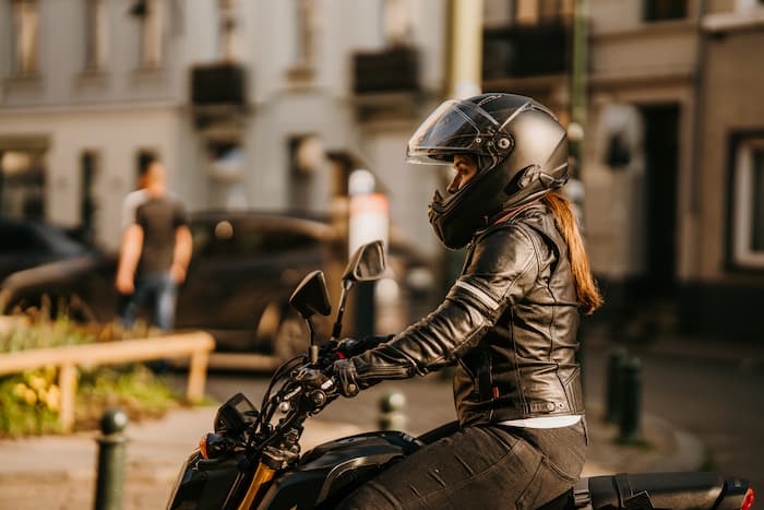 A female motorcyclist wearing a motorcycle helmet, jacket and gloves rides through the city.