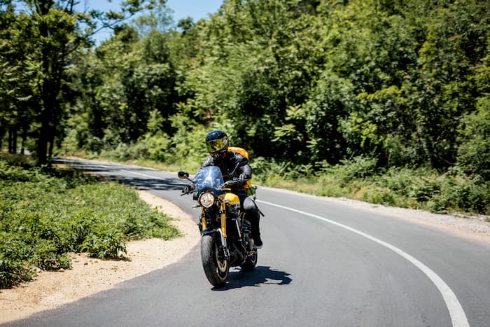 A motorcyclist rides a touring bike on a winding road surrounded by green trees and shrubs.