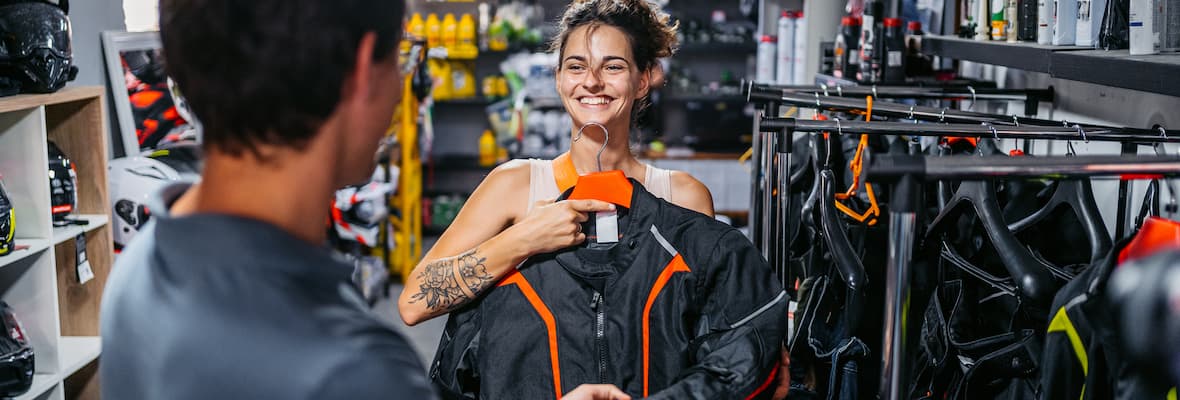 A young woman and her partner shop for a new motorcycle jacket in a store full of motorcycle gear.