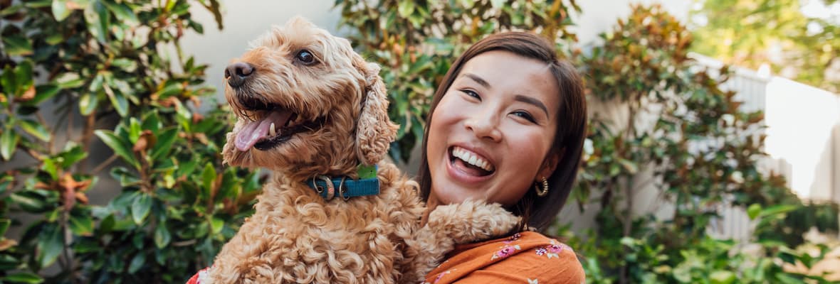 A happy young woman holds her golden cavoodle in a leafy green backyard