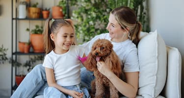 A mother and her young daughter sit with their pet miniature poodle on a couch in their outdoor patio.