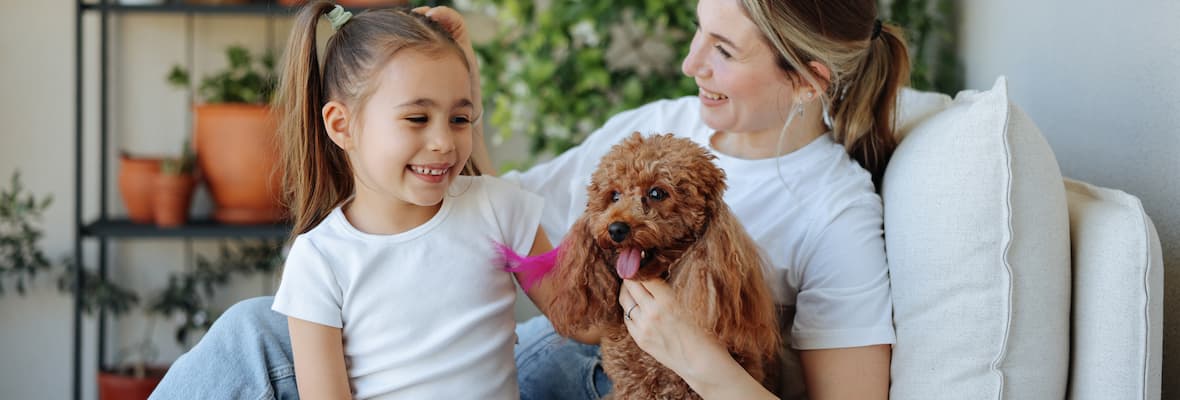 A mother and her young daughter sit with their pet miniature poodle on a couch in their outdoor patio.