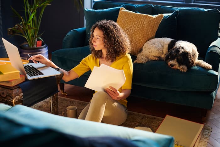 A young woman sits on her living room floor to use her laptop while her dog sleeps behind her on a couch.