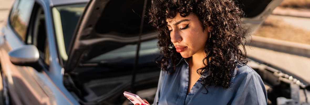 A young woman uses her mobile phone to contact Roadside Assistance in front of her broken-down car on the side of the road.